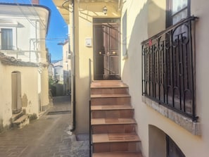 Une rue étroite avec un escalier menant à l’entrée d’un immeuble, une porte en métal et un ciel bleu dégagé.