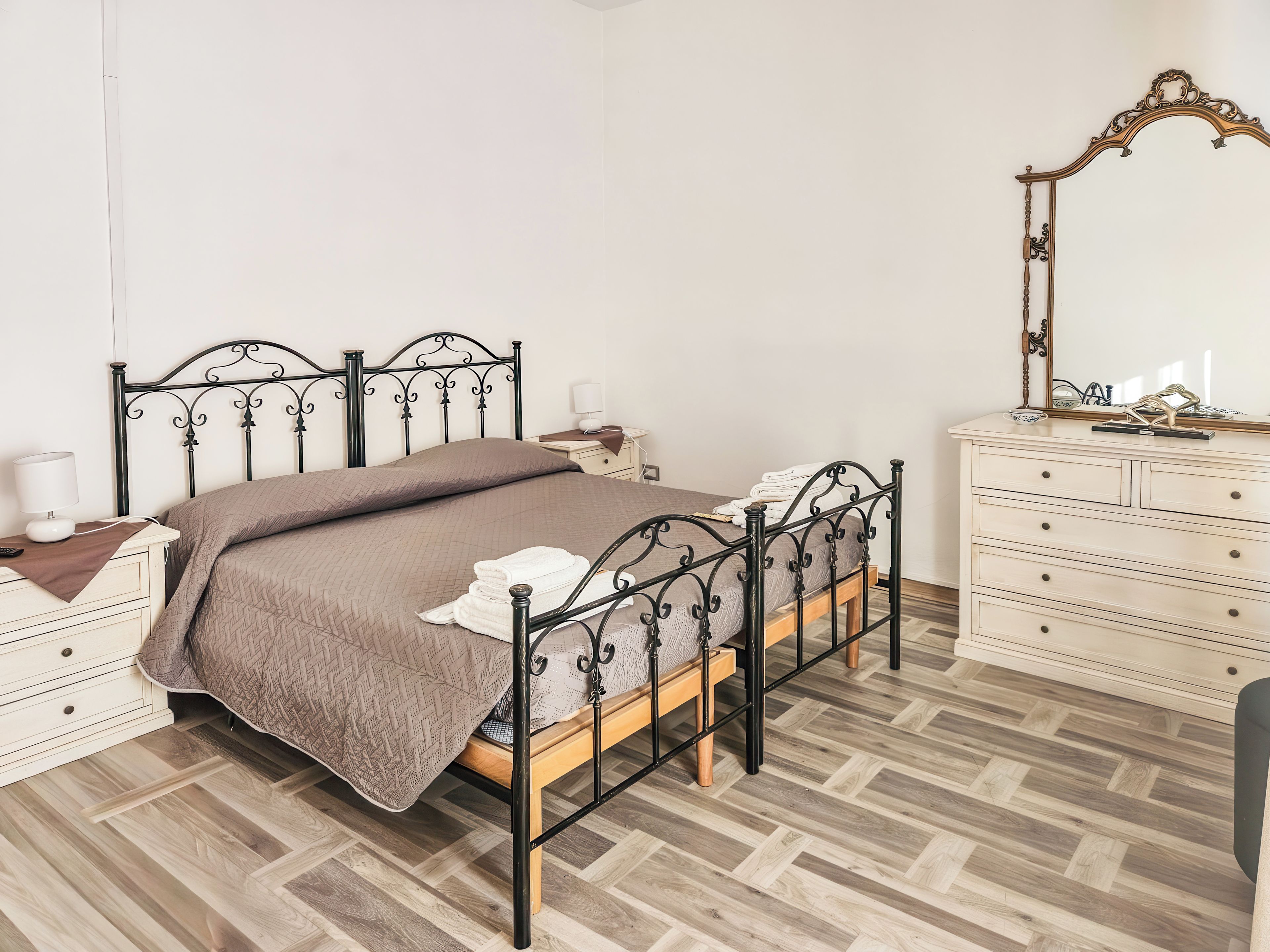 Une chambre à coucher bien rangée, avec un lit en métal, une table de chevet avec une lampe, une commode avec des tiroirs et un miroir accroché au mur.