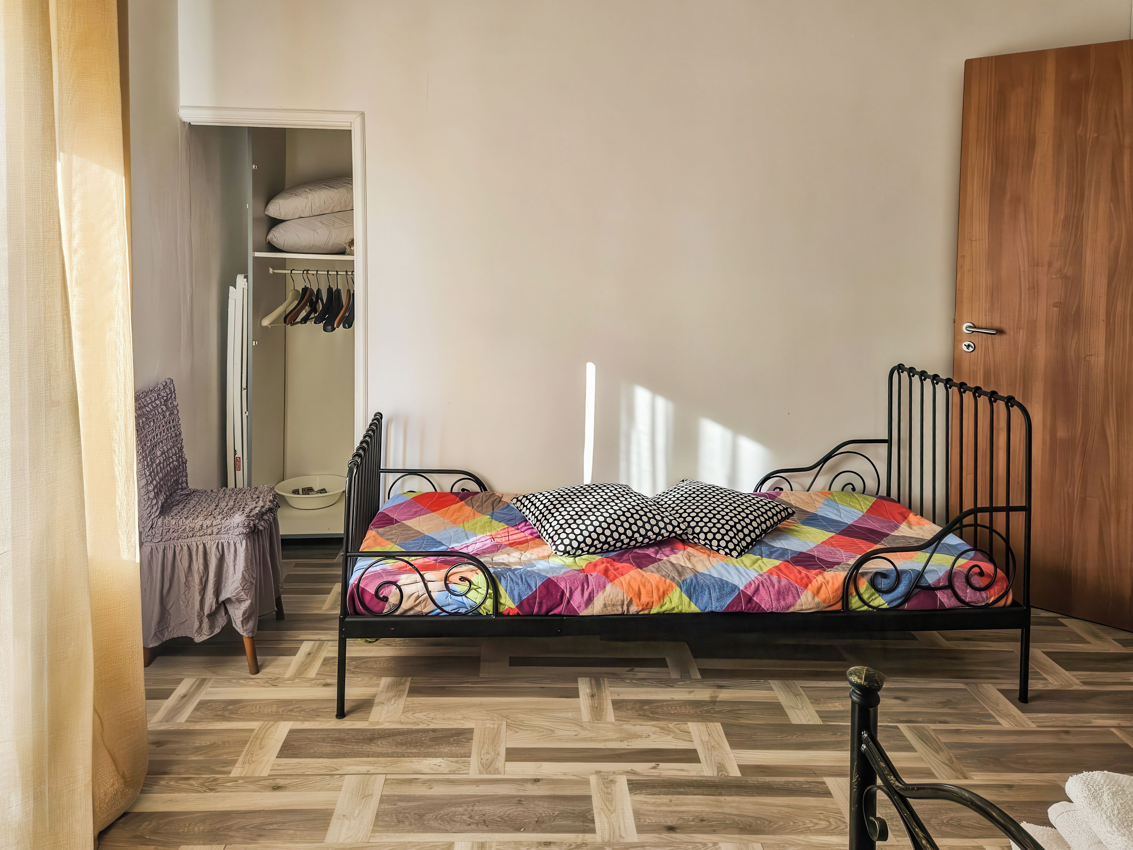 Una cama individual con colcha colorida, un armario de madera y un vestidor con ropa colgada.