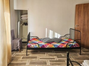 A single bed with a colorful quilt, a wooden wardrobe, and a closet with hanging clothes.