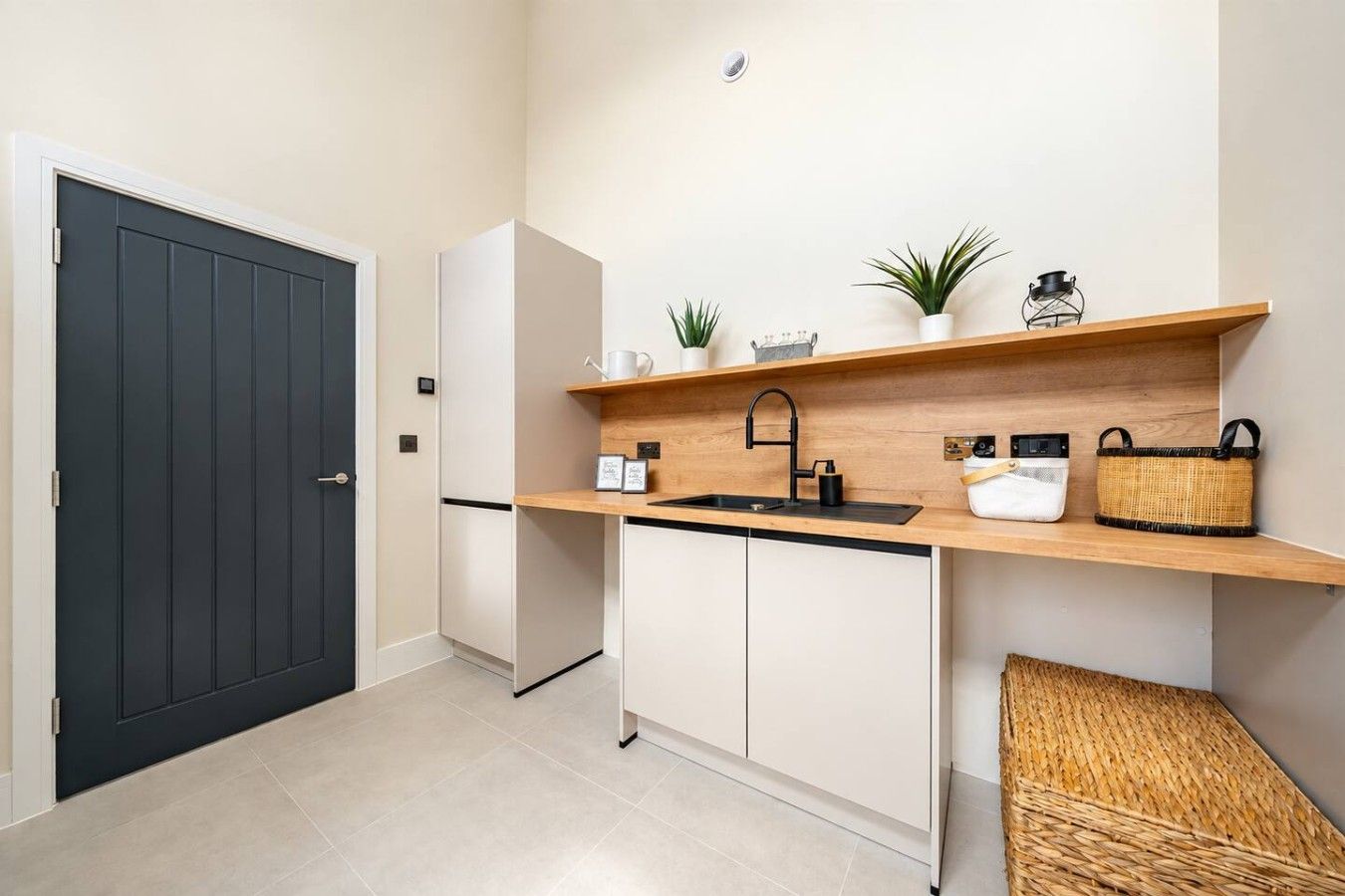 Una cucina compatta con una porta scura, pensili bianchi, un piano di lavoro in legno, un lavello nero e un piccolo ripiano con piante e un cesto.