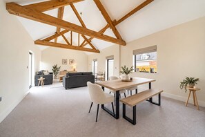 A spacious loft with exposed wooden beams, a dining area with a wooden table and benches, a grey sofa, and a small round table with a plant.
