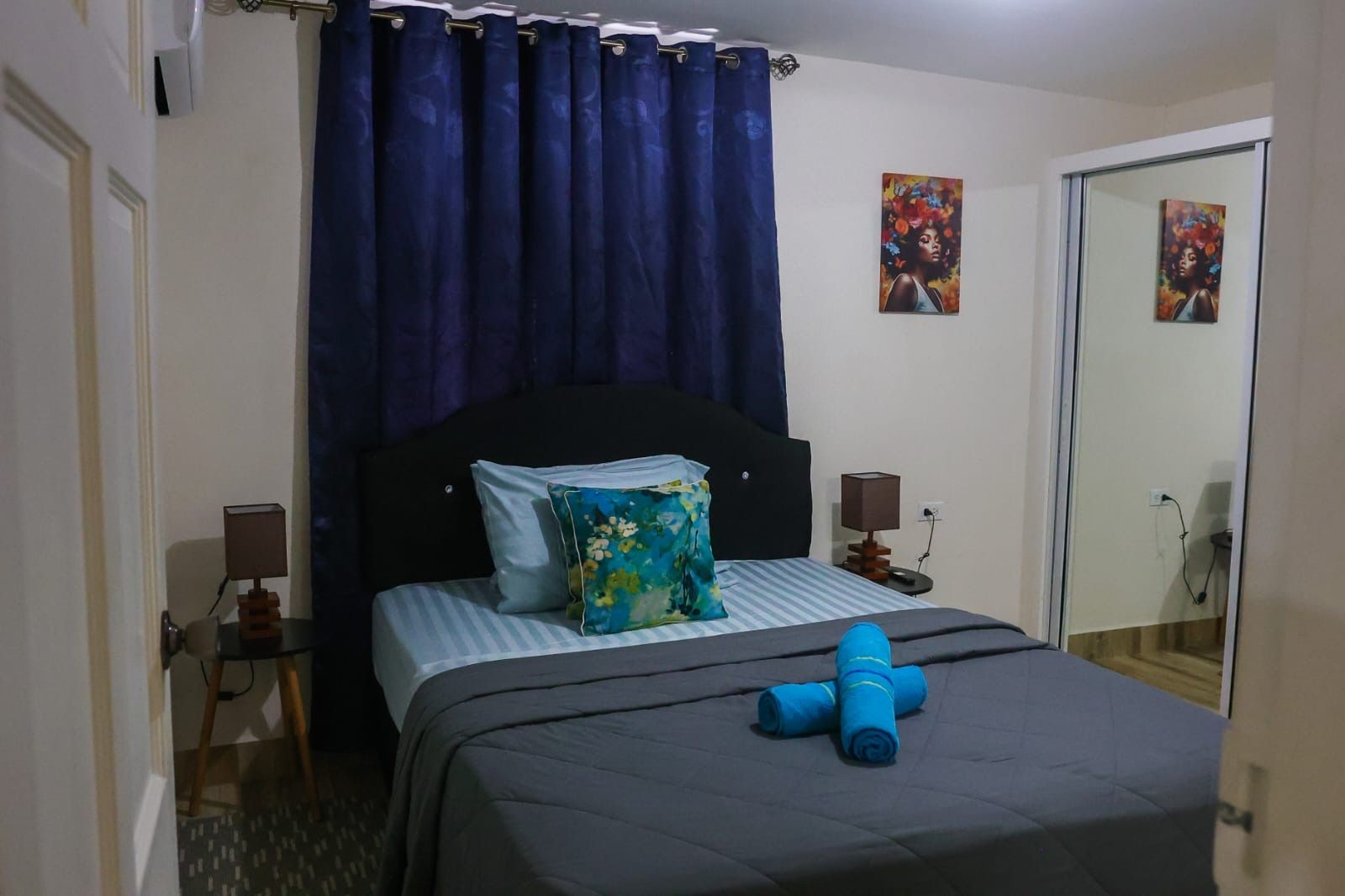 A hotel room with a bed, a blue curtain, a nightstand with a lamp, and two paintings on the wall.