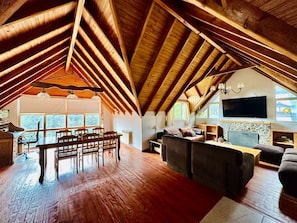 Een zolderkamer met een hoog houten plafond, een zithoek met stoelen en een tafel, een bank, een televisie en een stenen open haard.