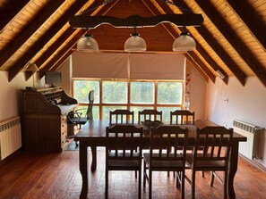 Een eetruimte met een houten tafel en stoelen, een raam met uitzicht op bomen, en een plafond met houten balken en hangende lampen.