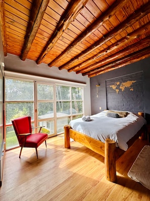 Een slaapkamer met een houten plafond, een groot raam, een bed met een wit matras en een rode stoel.