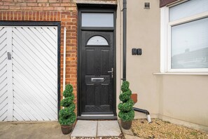 A black front door with a glass panel, flanked by two potted plants, and a white garage door to the left. - 10 mins from city centre. 6 Bed all ensuite, contractor friendly whole house (Peterborough)