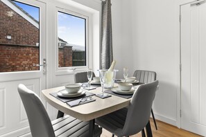 A dining area with a table set for four, chairs, a window with curtains, and a view of a brick building outside. - 10 mins from city centre. 6 Bed all ensuite, contractor friendly whole house (Peterborough)