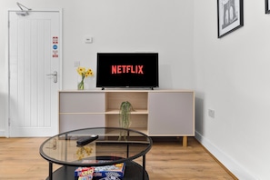 Uma sala de estar moderna com uma televisão exibindo 'NETFLIX', uma mesa de centro de vidro com um controle remoto, um vaso com flores e uma estante de livros.