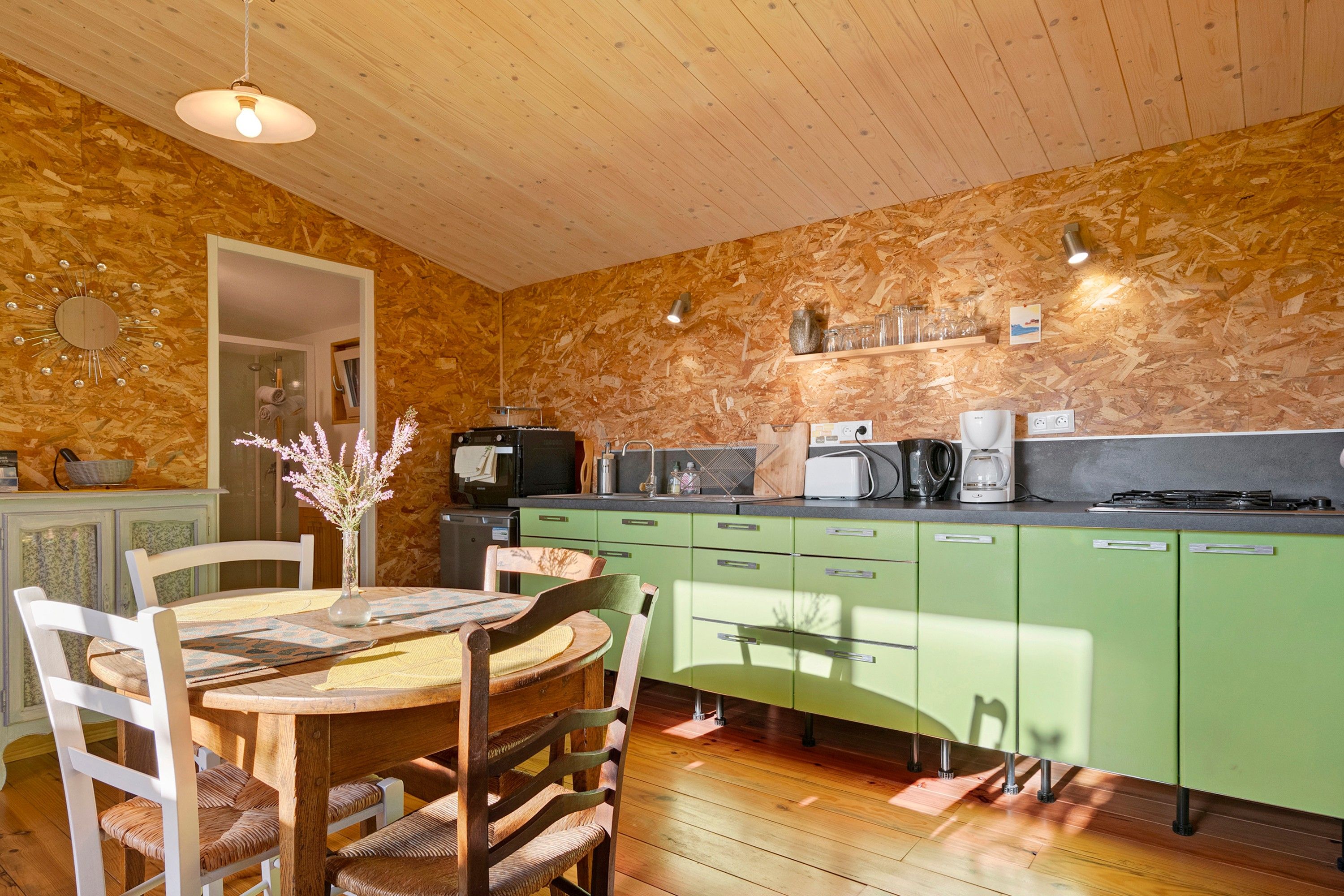A kitchen with a wooden ceiling, green cabinets, a dining table with chairs, and a vase with flowers.