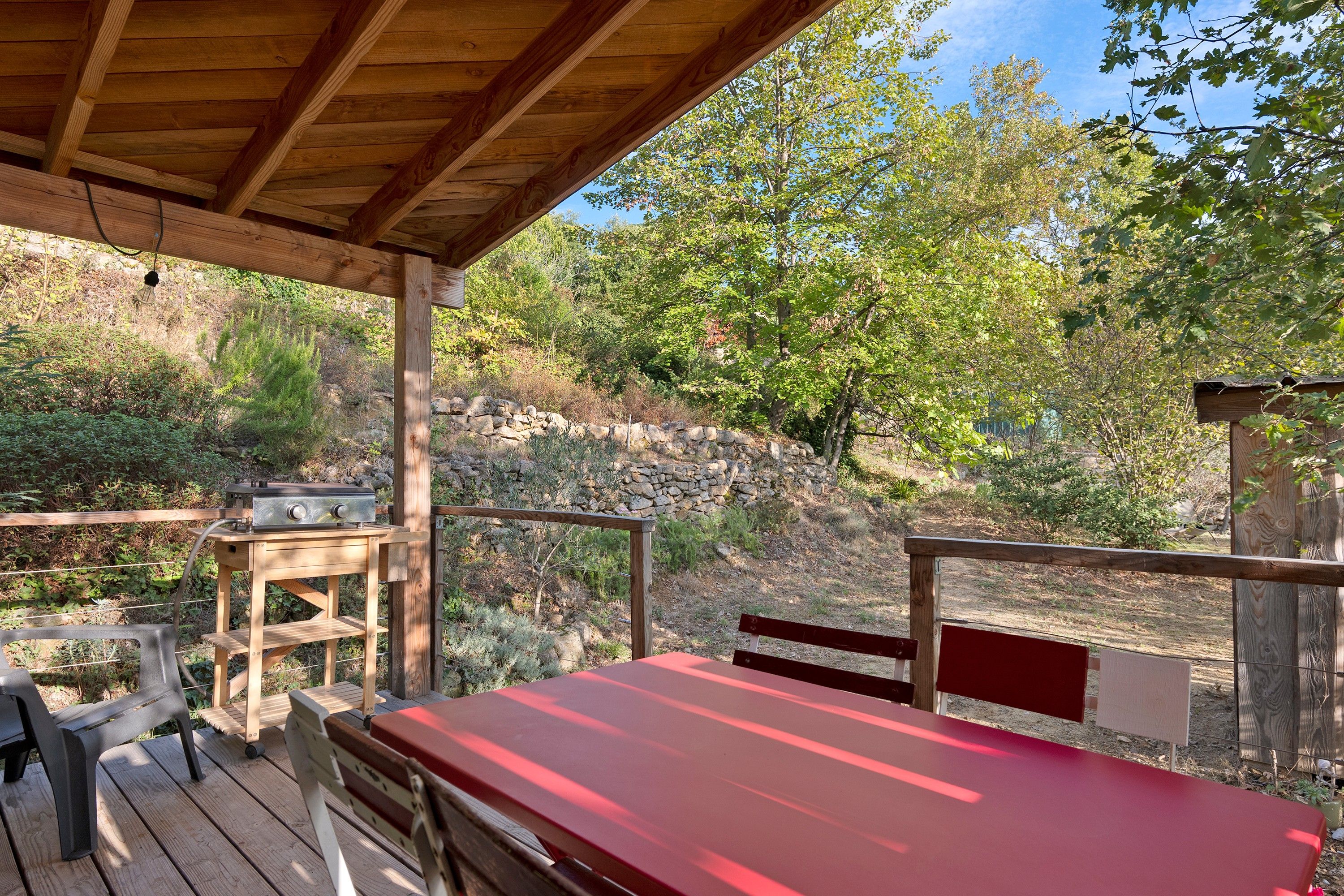 An outdoor patio with a red table and benches, a barbecue grill, and a stone wall in the background.