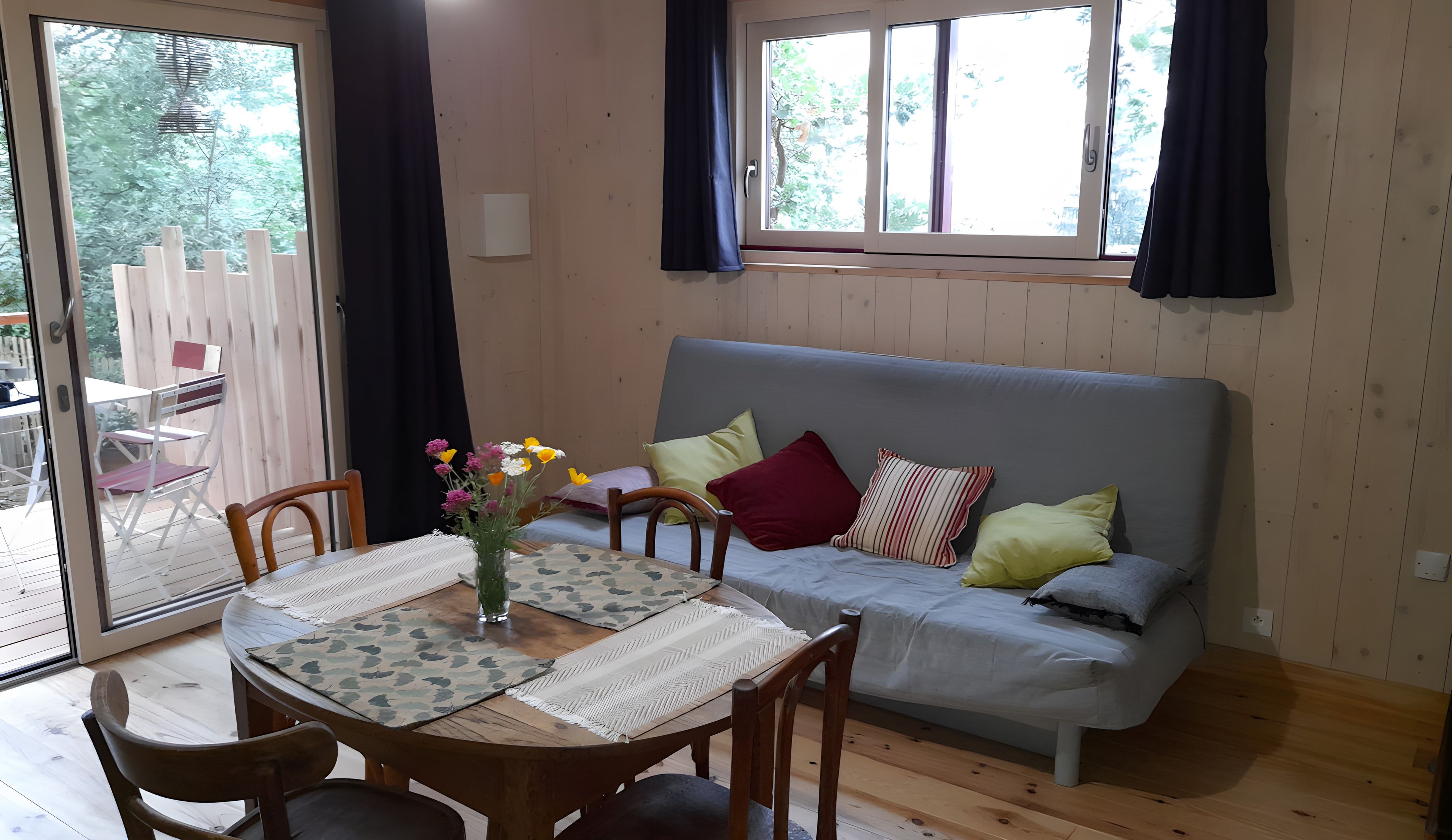 A room with a wooden floor, a grey sofa with cushions, a wooden dining table with chairs, and a window with curtains.