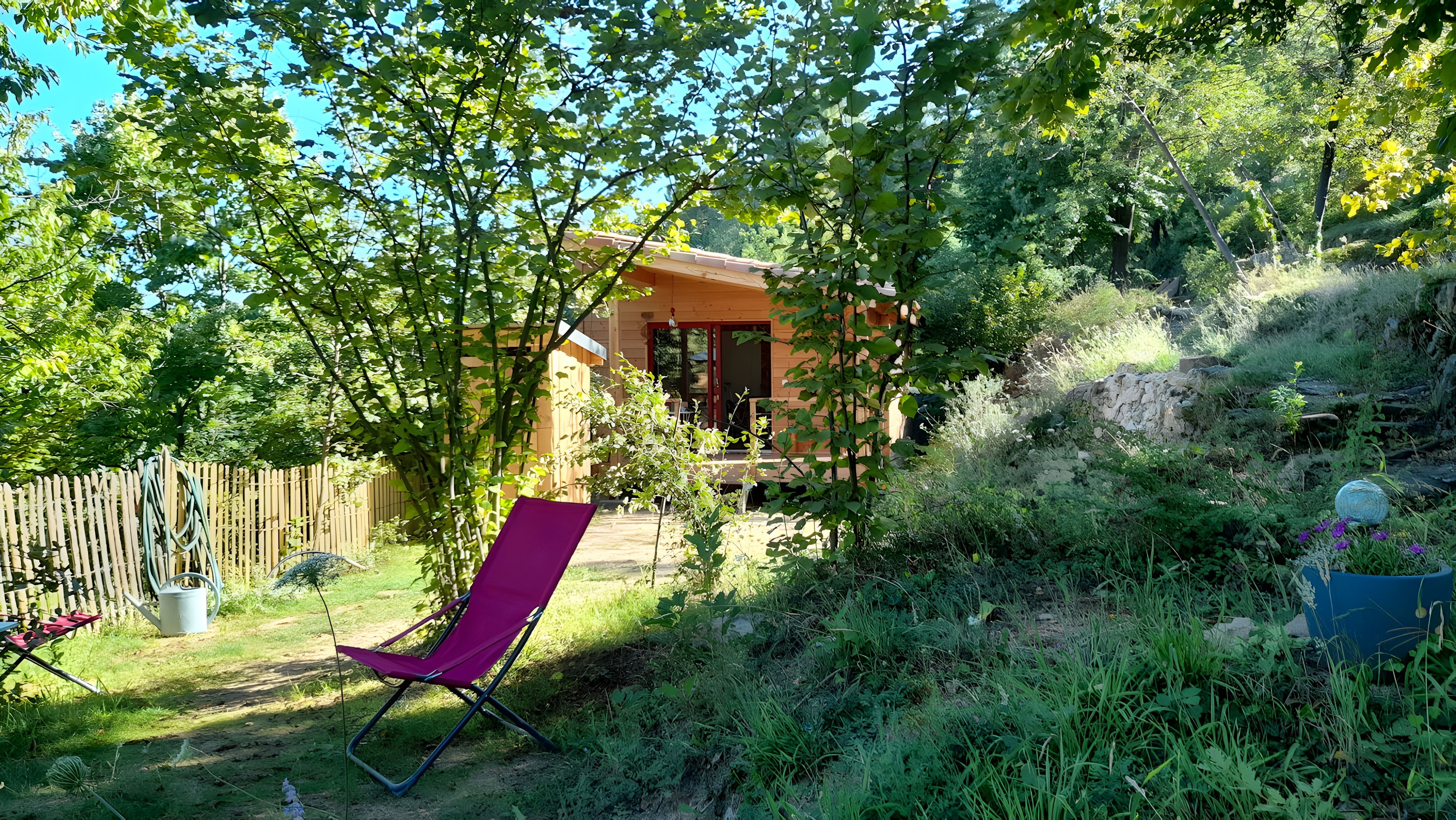 Une cabane en bois dotée d’un porche, entourée d’une végétation luxuriante et d’une clôture en bois.