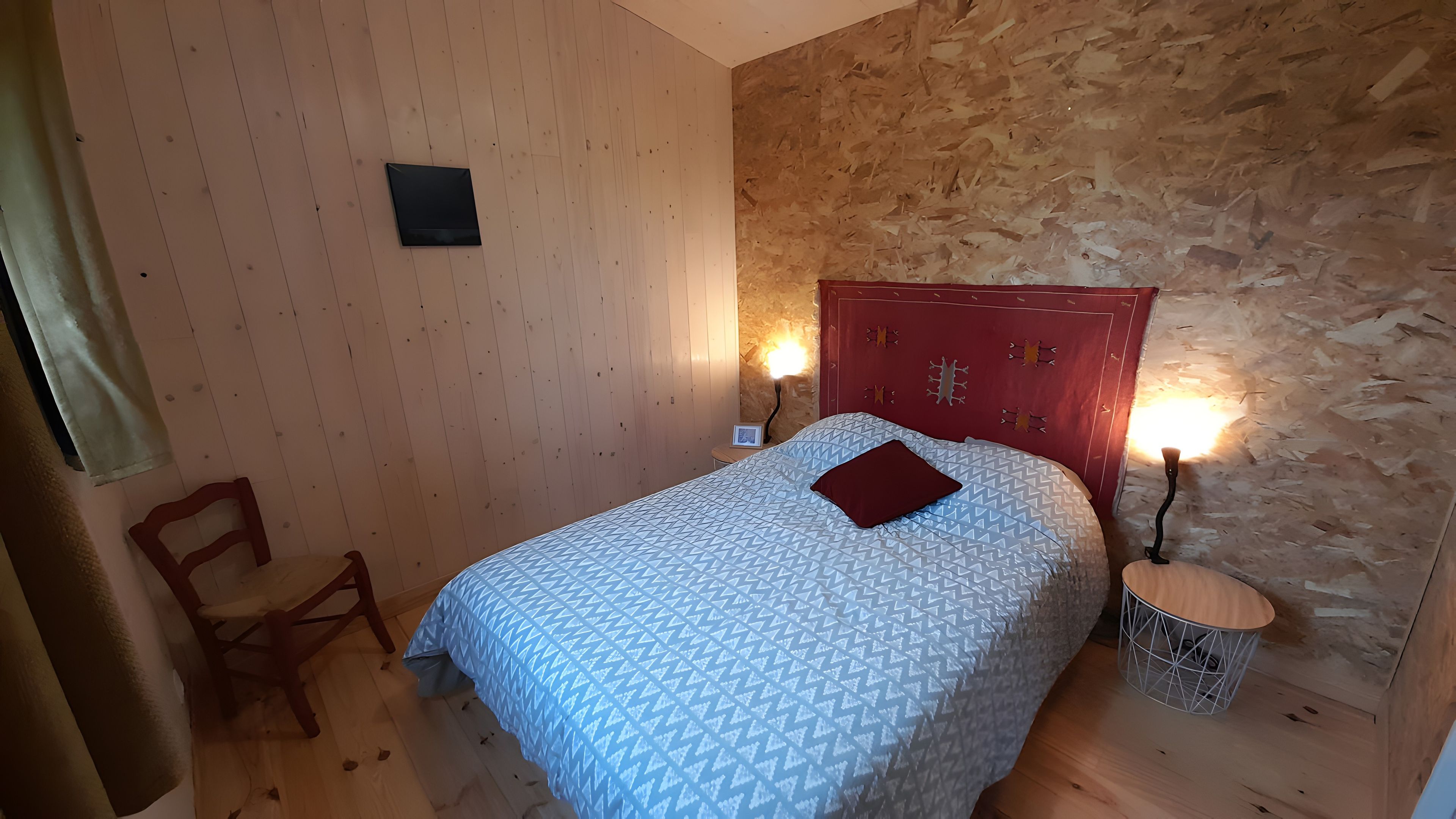 Une chambre à coucher avec une tête de lit en bois, un lit recouvert d’une literie à motifs, une chaise et une table de chevet avec une lampe.