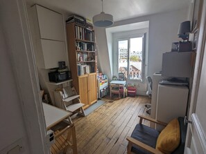 A compact living space with a kitchenette, a wooden bookshelf, a dining area with a small table and chairs, and a view through a large window. - Charming 40m² apart for 2 people – Paris 10 (Paris)