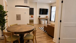 A dining area with a round wooden table and chairs, a television mounted on the wall, and a window with curtains. - The James Suite Three | New 2BR Beach Block (Atlantic City)