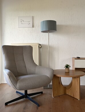 A modern room with a grey upholstered armchair, a wooden side table with a plant, a floor lamp, and a radiator. - Cozy vacation rental in Oberfrauenwald near Waldkirchen with a magnificent panoramic view (Waldkirchen)