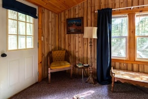 Ein Zimmer mit Holzwänden, einer Tür, einem Fenster mit Vorhängen, einem Stuhl, einem kleinen Tisch mit Lampe und einer Bank.