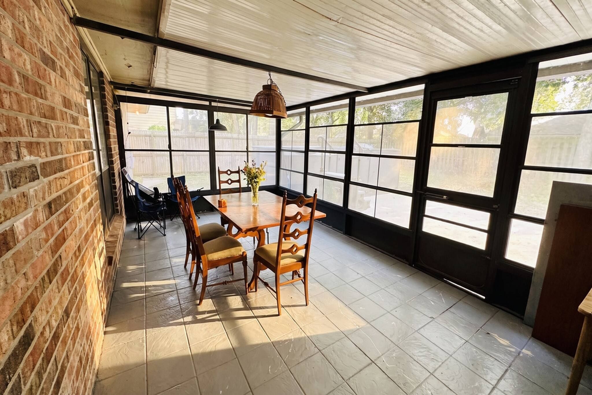 A sunlit patio with a wooden table and chairs, large windows, and a brick wall.