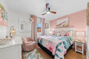 Ein Schlafzimmer mit Bett, Stuhl, Nachttisch, Deckenventilator und einem Fenster mit Vorhängen.