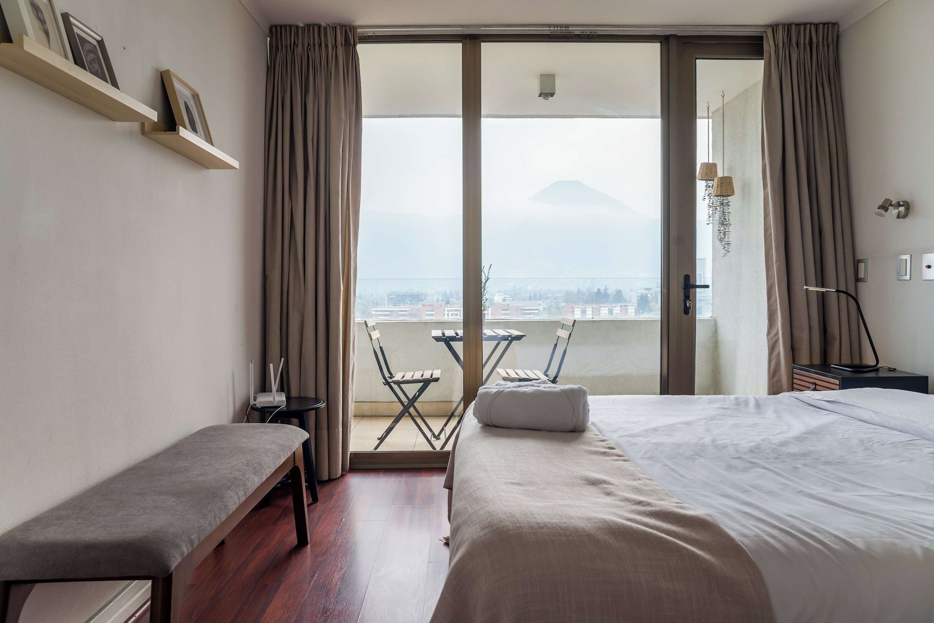 A hotel room with a bed, a bench, a small table with a lamp, a chair, and a balcony with a view of a mountain.