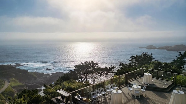 A seaside terrace with tables and chairs, overlooking the ocean and distant landforms.