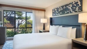 Ein Hotelzimmer mit einem großen Bett, einem Nachttisch, einer Lampe, einem Gemälde und Blick auf Bäume durch ein Fenster.
