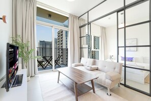 A modern living room with a sofa, a coffee table, and a TV on a stand. There is a balcony with a view of high-rise buildings. - Modern 1BR Retreat in Socio | Dubai Hills (Dubai)