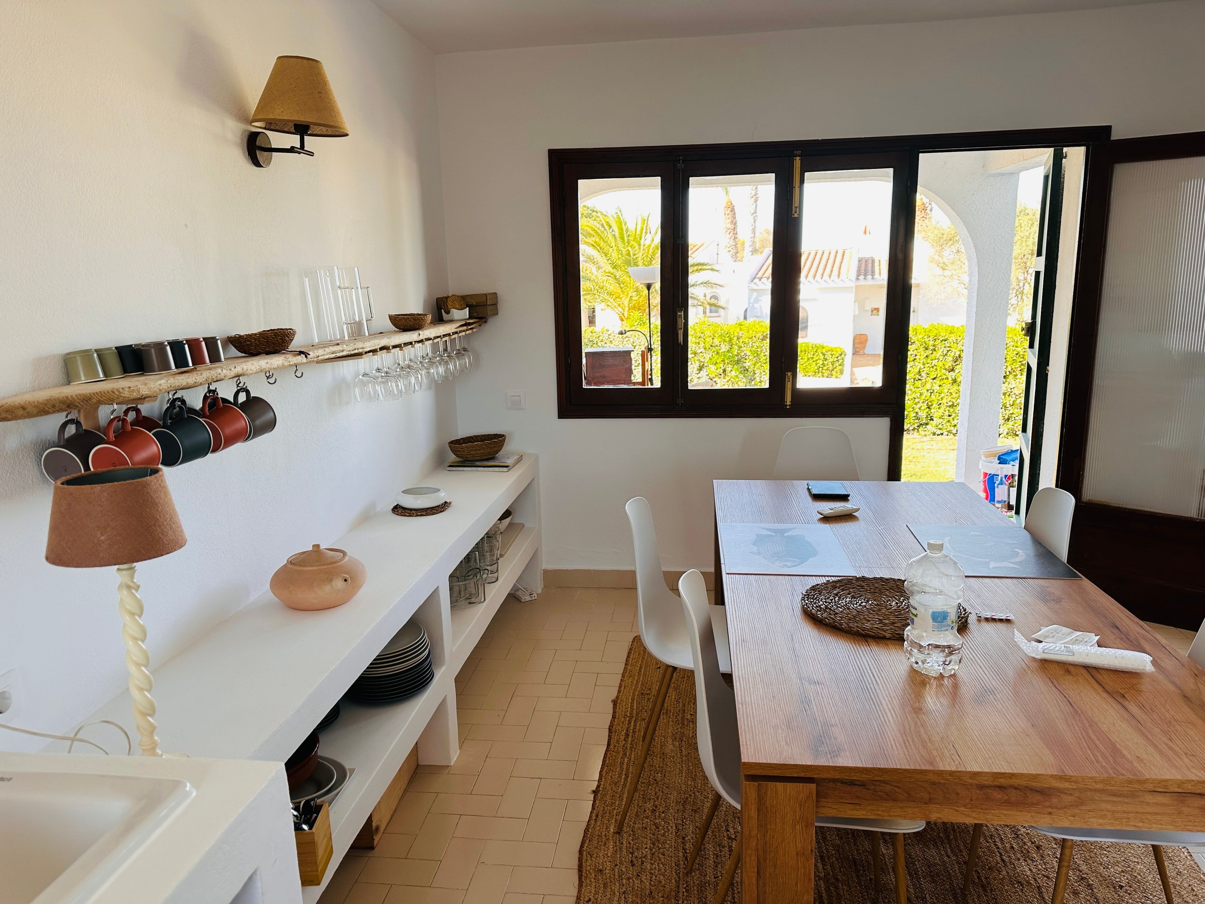 Un comedor con una mesa de madera, sillas, un estante blanco con utensilios de cocina y una ventana con vistas al follaje.