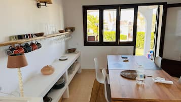 Un comedor con una mesa de madera, sillas, un estante blanco con utensilios de cocina y una ventana con vistas al follaje.