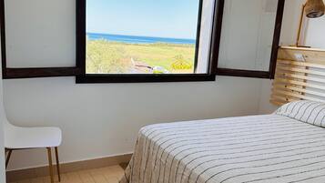 A bedroom with a large window, a bed with a striped blanket, a white chair, and a wooden bedside table with a lamp.