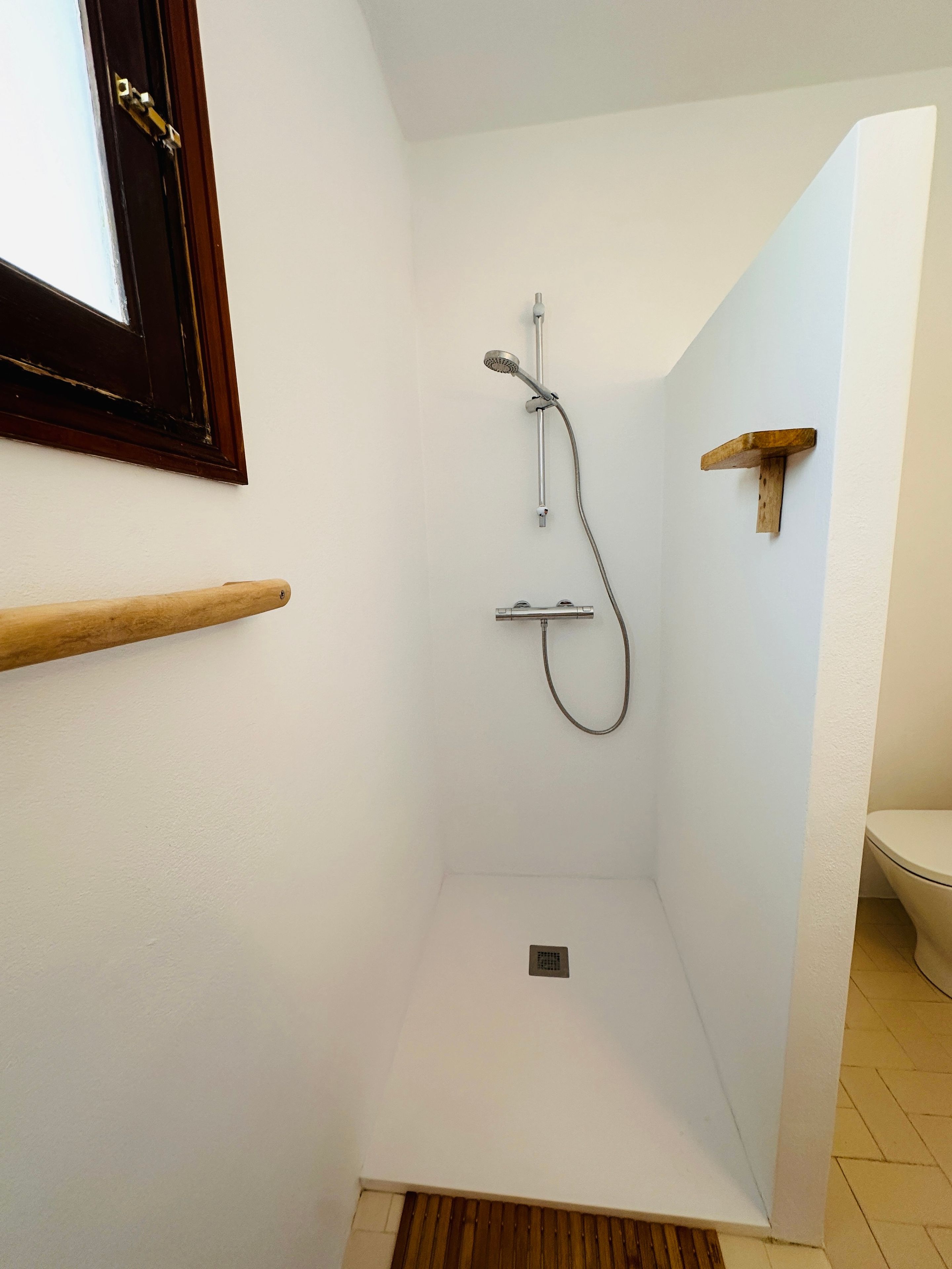 A small, enclosed shower area with a showerhead and hose, a wooden shelf, and a toilet visible in the corner.