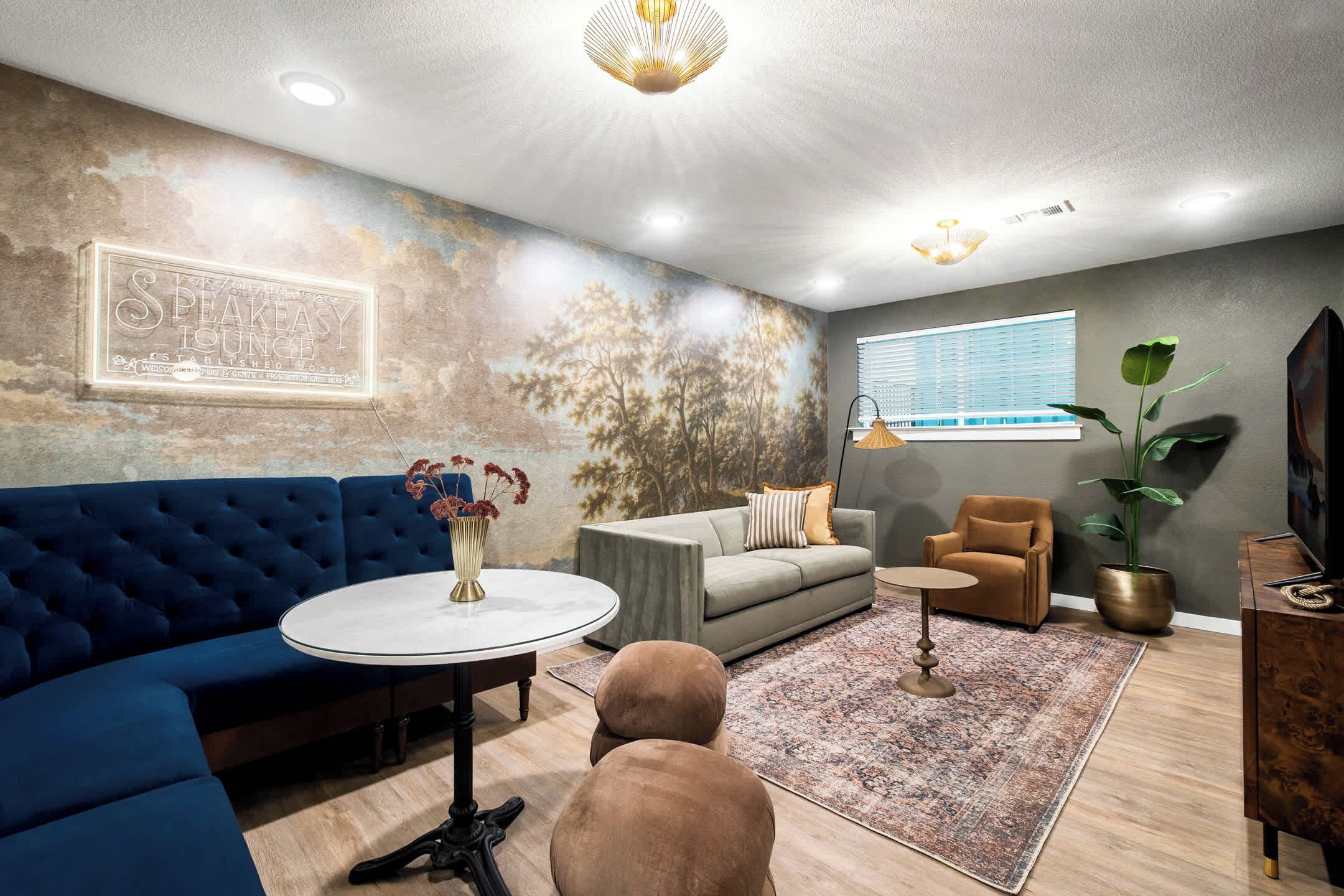 Ein modernes Wohnzimmer mit einem blauen Sofa, einem runden Couchtisch und einem grauen Sofa. An der Wand hängt ein Gemälde und es gibt ein Fenster mit Jalousien.
