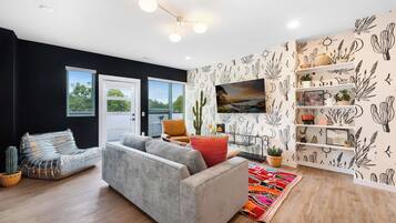 Ein modernes Wohnzimmer mit einem grauen Sofa, einem Couchtisch und einem farbenfrohen Teppich. Eine schwarze Wand zeigt ein Design mit Kakteen und Sonne, und ein Fenster mit Jalousien.