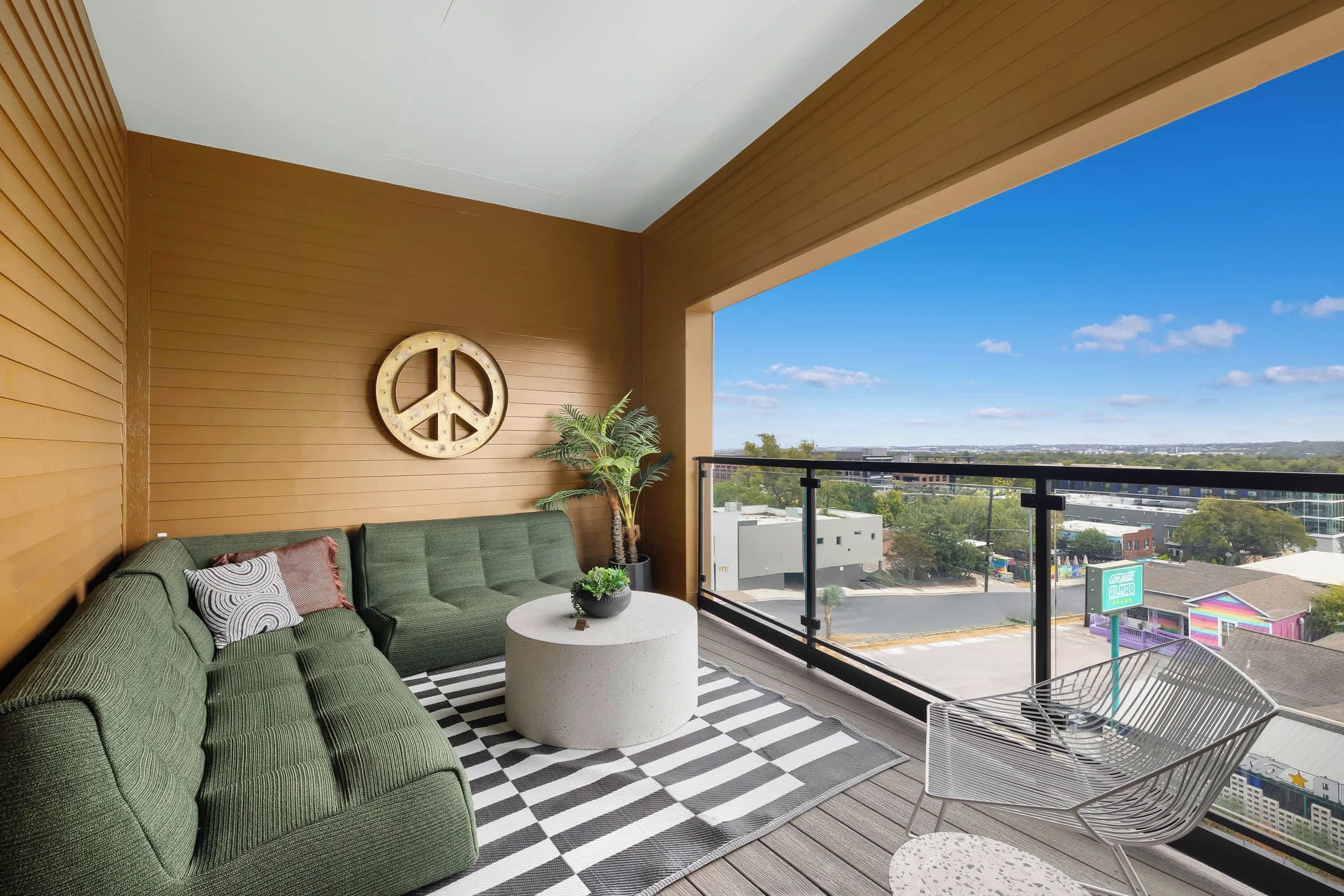 Ein Balkon mit einer grünen Couch, einem runden Couchtisch und einem Metallstuhl. An der Wand hängt eine Friedenssymbol-Dekoration, und es gibt einen Blick auf Gebäude und Grünflächen.