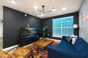 Una sala de estar moderna con un televisor de pantalla plana grande, un sofá azul, una mesa de centro y un amplio ventanal con persianas.
