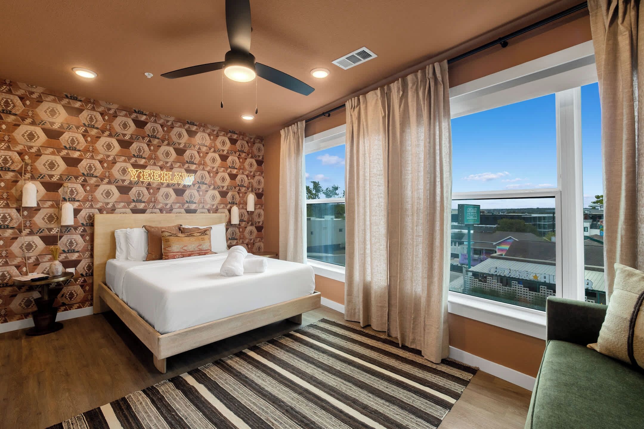 Ein Hotelzimmer mit einem Bett, einem Deckenventilator, Blick auf die Stadt und einer Wand mit Muster.
