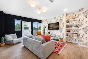 Ein modernes Wohnzimmer mit einem grauen Sofa, einem Couchtisch und einem farbenfrohen Teppich. Eine schwarze Wand zeigt ein Design mit Kakteen und Sonne, und ein Fenster mit Jalousien.