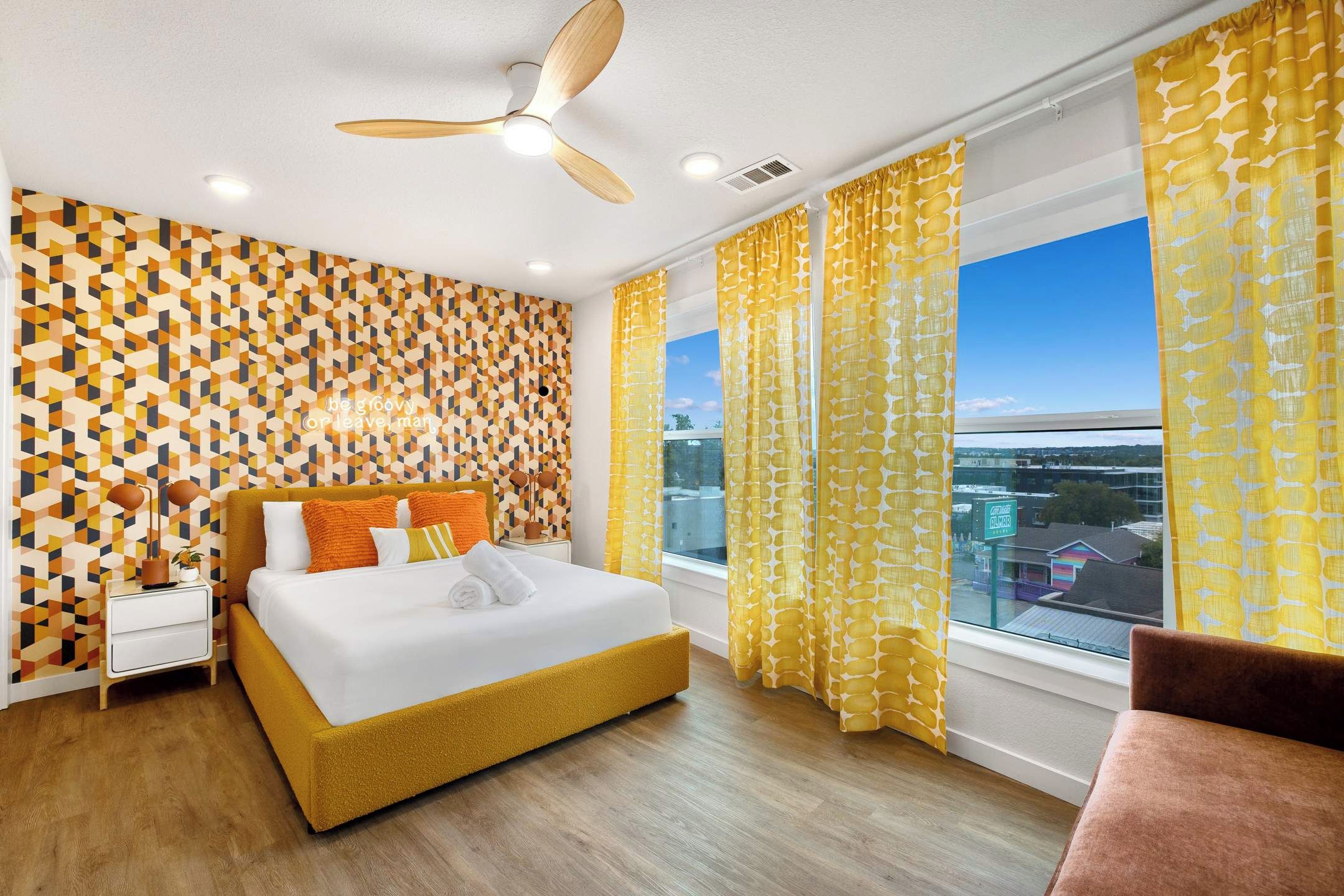 Ein modernes Schlafzimmer mit Wand in geometrischem Muster, einem Bett mit orangefarbener Bettwäsche, einem Deckenventilator und Blick auf eine Stadtlandschaft durch große Fenster.