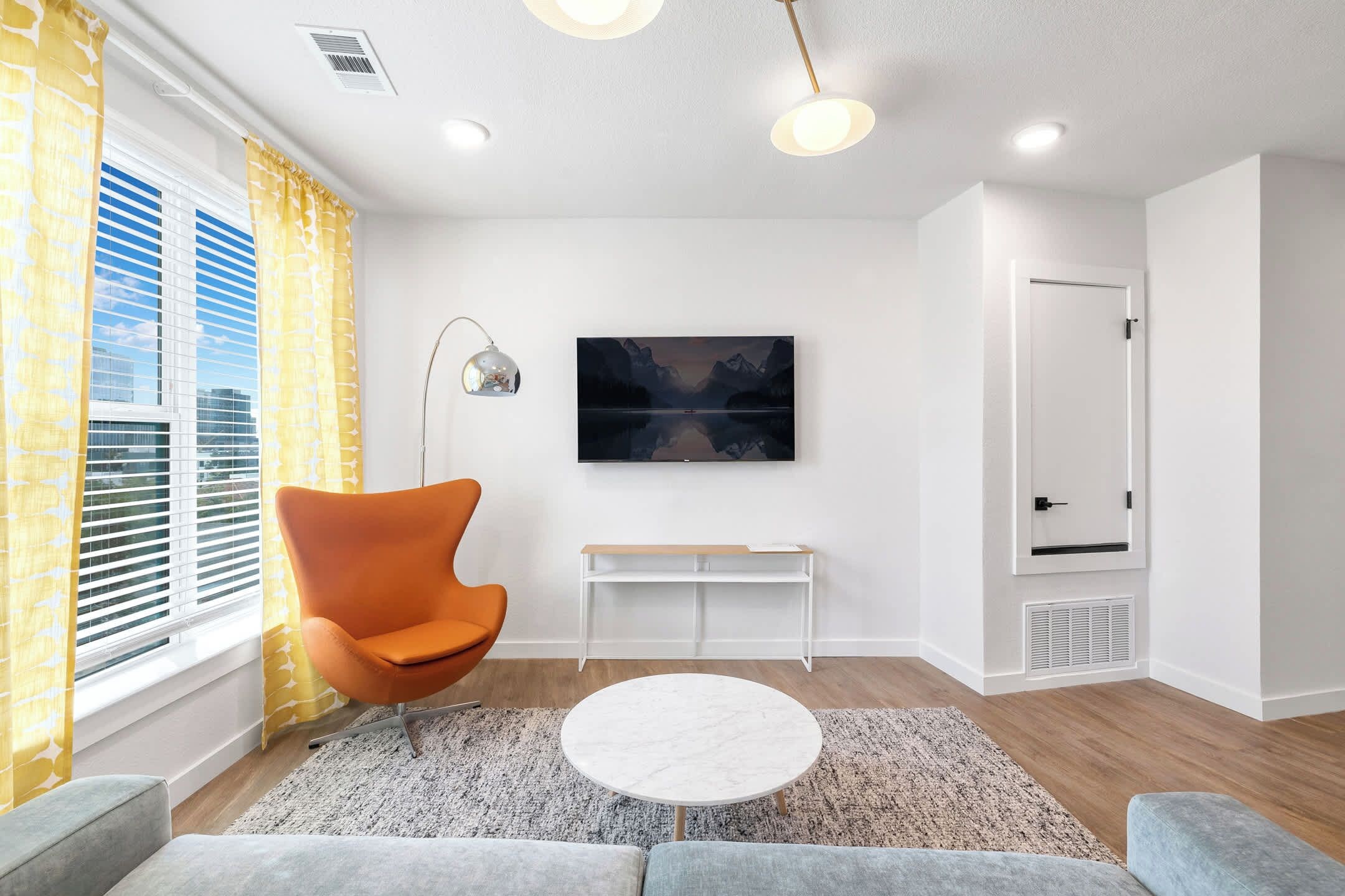 Ein modernes Wohnzimmer mit einem großen Flachbildfernseher, einem einzelnen orangefarbenen Sessel, einem runden Couchtisch und Blick auf die Stadt durch große Fenster.