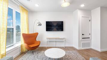 Ein modernes Wohnzimmer mit einem großen Flachbildfernseher, einem einzelnen orangefarbenen Sessel, einem runden Couchtisch und Blick auf die Stadt durch große Fenster.