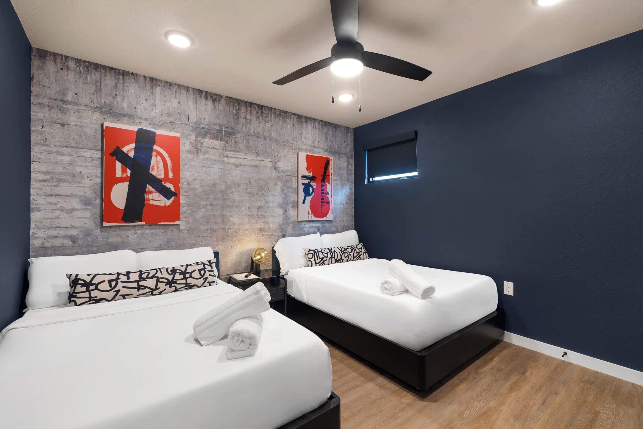 Ein Hotelzimmer mit zwei Betten, einem Deckenventilator, Kunstwerken an der Wand und einem Fenster.