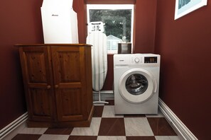A laundry room with a washing machine, a wooden cabinet, and a window with a view of a house. - Central Warrington 2-Bed Home near Hospital & Train Station | Free Parking (Warrington)
