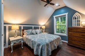 Ein Schlafzimmer mit Bett, Nachttischen, einem Schminktisch, einem Deckenventilator und einem Fenster mit Blick auf Bäume.