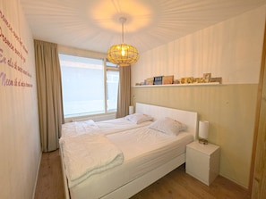 Ein Schlafzimmer mit einem großen Bett, einem Nachttisch, einer Hängeleuchte, einem Fenster mit Vorhängen und einer Wand mit einem Regal.