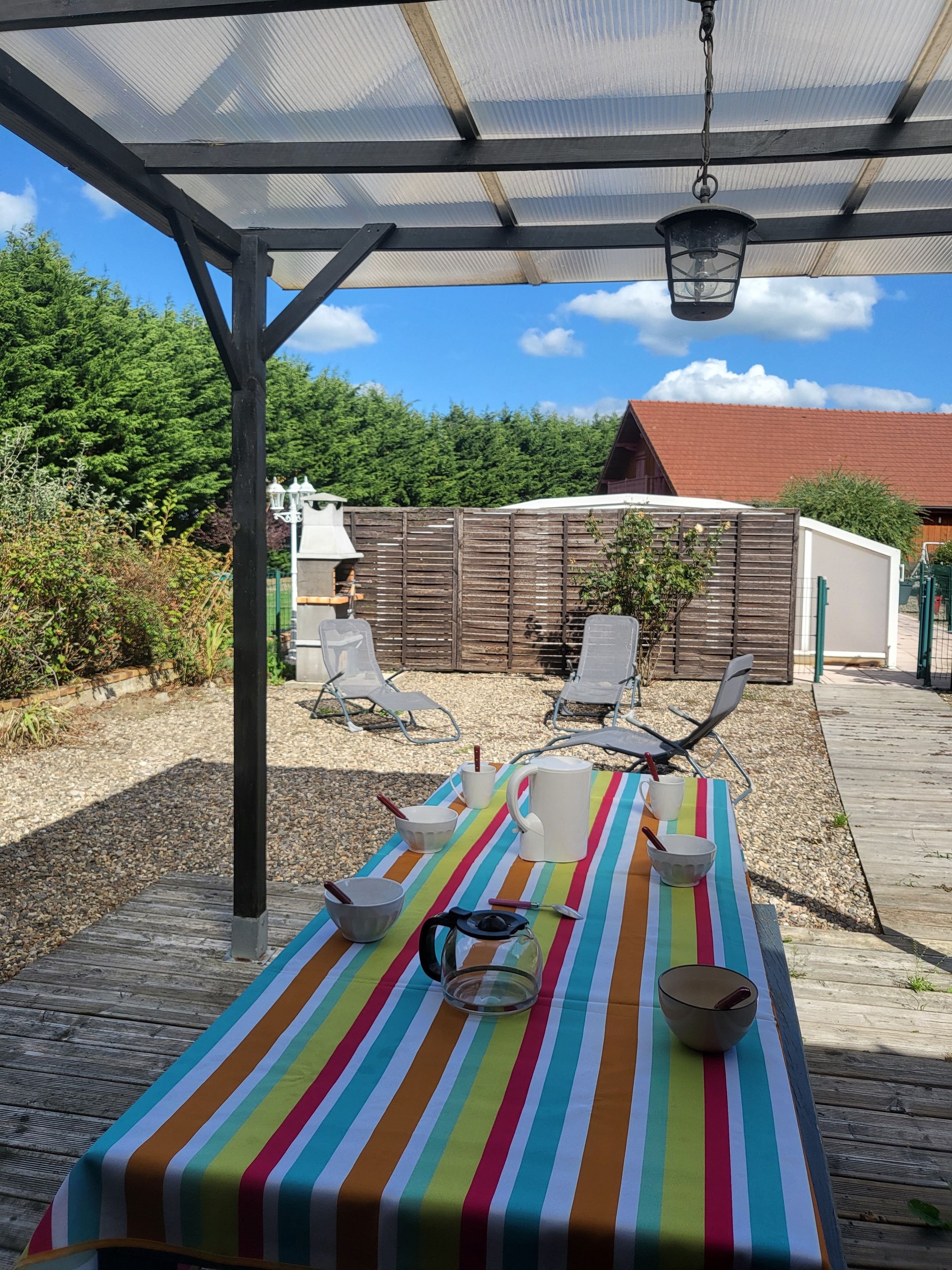 Une terrasse extérieure avec une table dressée pour le repas, entourée de chaises, située sous une zone abritée d’où l’on peut admirer une clôture en bois et de la verdure.