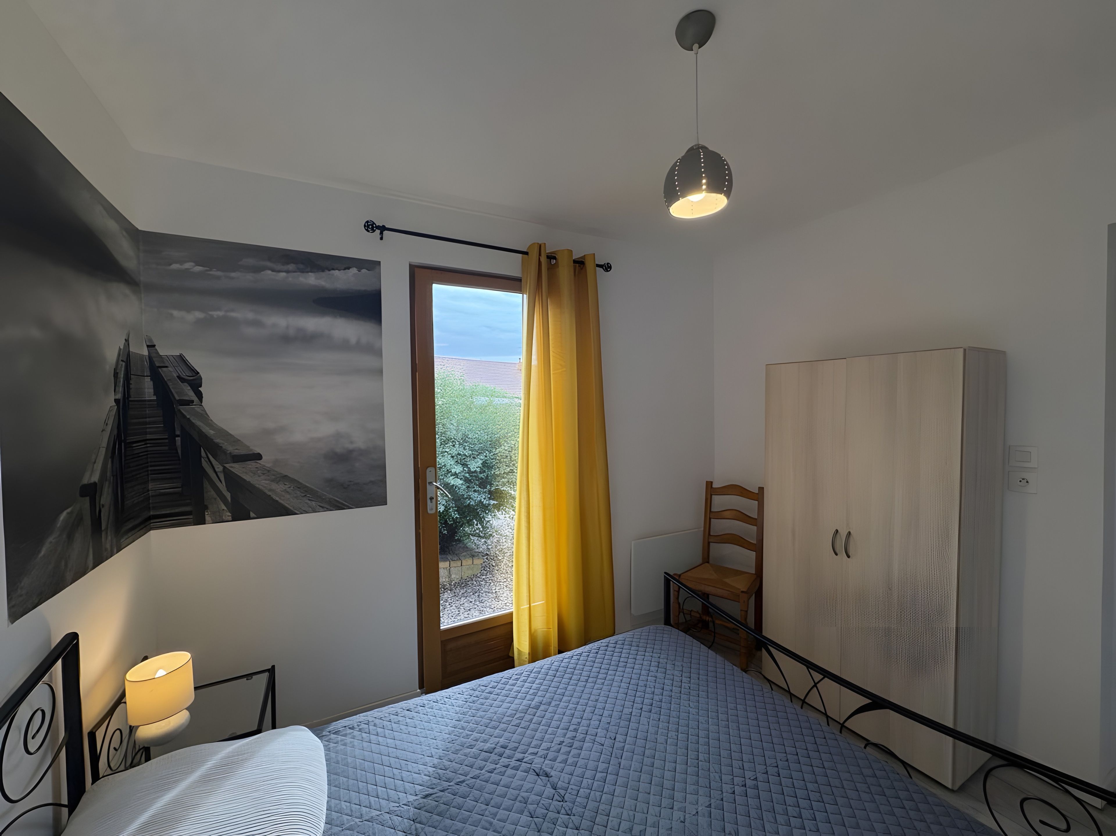 Een slaapkamer met een bed, nachtkastje, kledingkast, raam met gordijnen en een schilderij aan de muur.
