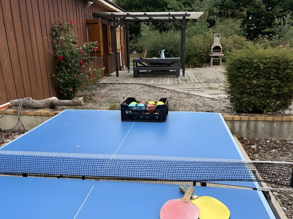 A ping pong table with a net, a black crate with colorful balls, and a wooden bench in a garden setting.