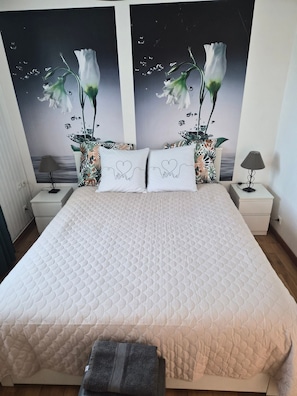 Cama bem arrumada com cobertura acolchoada, duas mesas de cabeceira com abajures e uma parede com uma pintura floral.