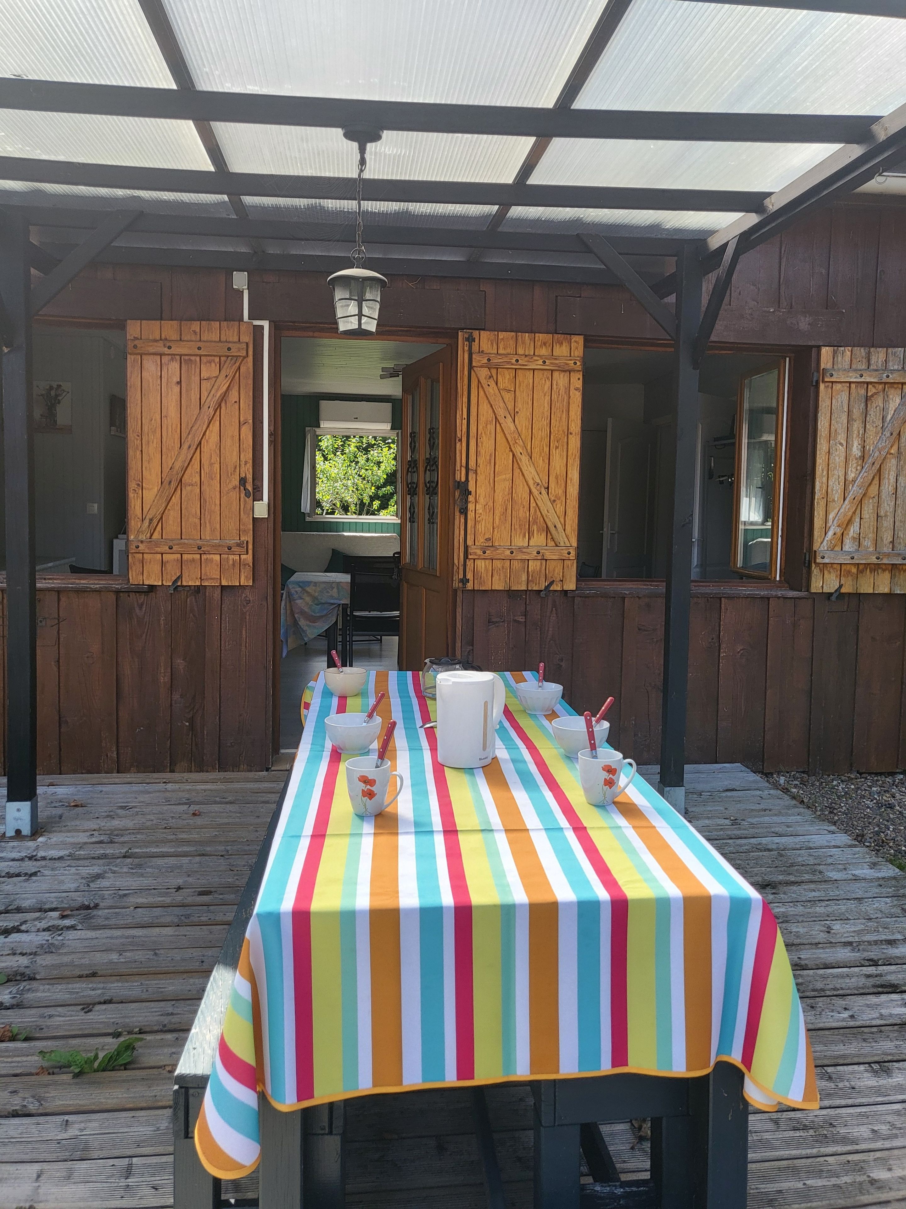 Een overdekt terras met een eettafel voor vier personen, met een geruit tafelkleed en witte bekers met rode handgrepen.